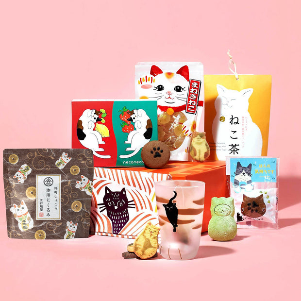 The Cat Lover's Box by Bokksu includes Japanese snacks and tea, cat-themed packaging, Manekineko Jelly, mini cat figurines, and a black cat glass, all beautifully arranged on a pink background.