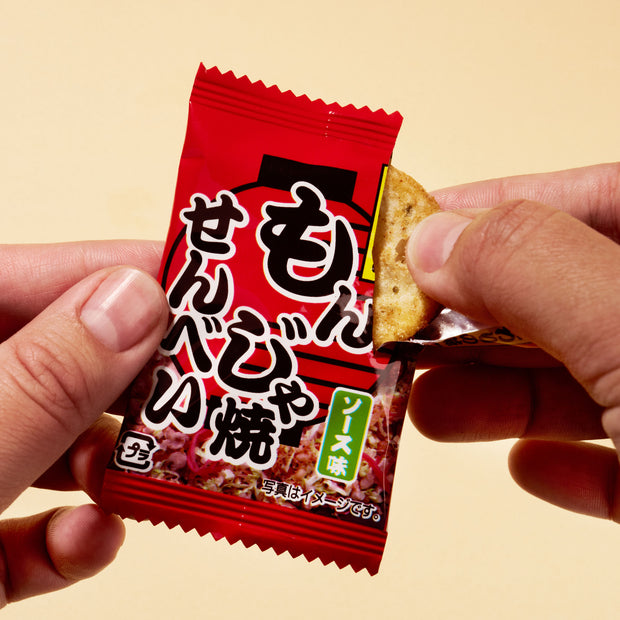 A person opens a red April ’26 Bokksu: Handpicked in Japan package of Japanese rice crackers, holding one cracker partially out of the wrapper against a beige background.