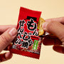 A person opens a red April ’26 Bokksu: Handpicked in Japan package of Japanese rice crackers, holding one cracker partially out of the wrapper against a beige background. thumbnail 13