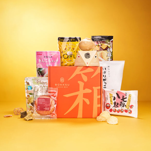 An assortment of Japanese snacks and instant soups surrounds an orange box labeled "The Japanese Savory Snack Box," featuring chips, cookies, crackers, and candy in vibrant packaging.
