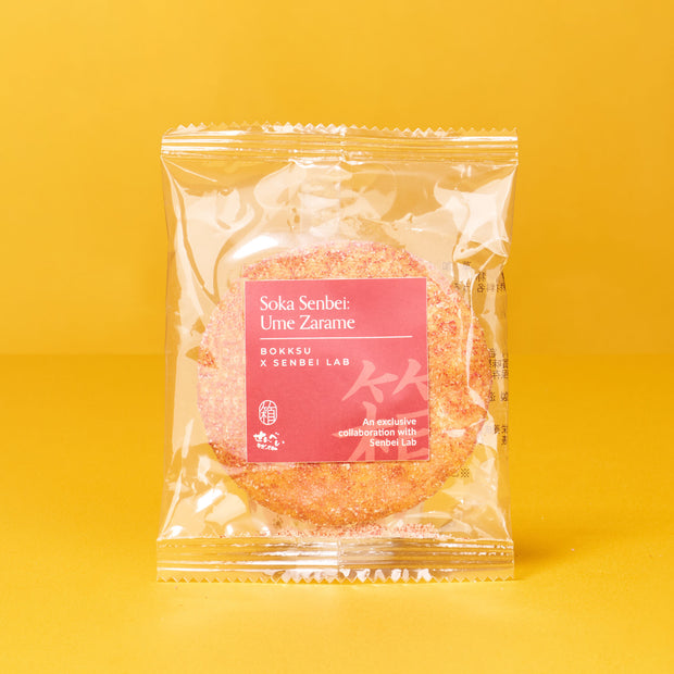A packaged Soka Senbei: Ume Zarame rice cracker from The Japanese Savory Snack Box rests on a yellow background, featuring a pink label with product and collaboration details.