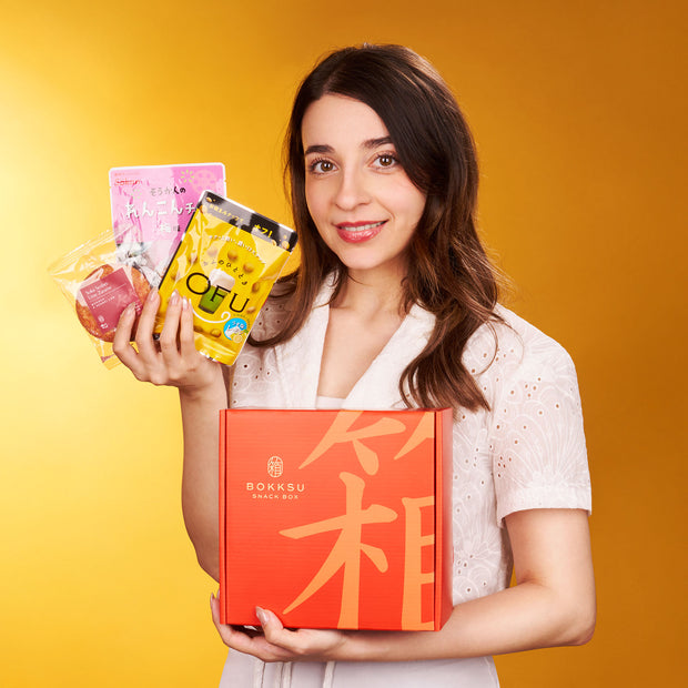 A woman in a white shirt holds The Japanese Savory Snack Box and several packaged Japanese snacks against a solid orange background.