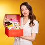 A woman in a white blouse holds an open The Japanese Savory Snack Box filled with assorted snacks against a yellow background. thumbnail 2