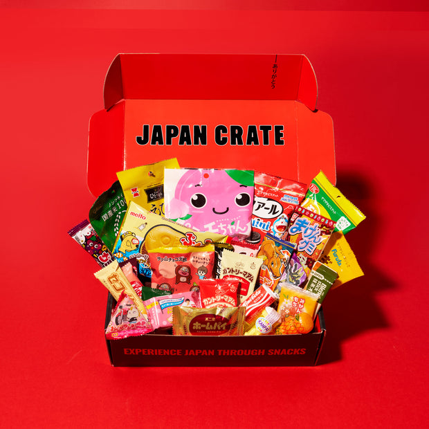 An open Dagashi Snack Crate (FBA) - Japanese Snack Variety Box with Candy, Chocolate, Cookies, Mochi & Ramune Treats is displayed on a vibrant red surface, filled with an assortment of colorful Japanese snacks and candies.
