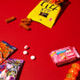 A selection of Japanese snacks and three white tablets are displayed on a red surface, with a yellow bag spilling out treats alongside vibrant candy packages—just like those you’d discover in the Dagashi Snack Crate (FBA) - Japanese Snack Variety Box with Candy, Chocolate, Cookies, Mochi &amp; Ramune Treats. thumbnail 3