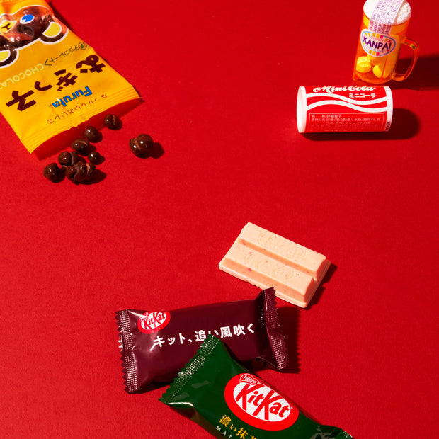 A Dagashi Snack Crate (FBA) – Japanese Snack Variety Box with candy, chocolate, cookies, mochi, and Ramune treats is displayed on a solid red background, featuring assorted Japanese snacks and candies such as yellow- and red-packaged treats, chocolate-coated beans, a small drink, and KitKat bars.