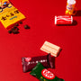 A Dagashi Snack Crate (FBA) – Japanese Snack Variety Box with candy, chocolate, cookies, mochi, and Ramune treats is displayed on a solid red background, featuring assorted Japanese snacks and candies such as yellow- and red-packaged treats, chocolate-coated beans, a small drink, and KitKat bars. thumbnail 4