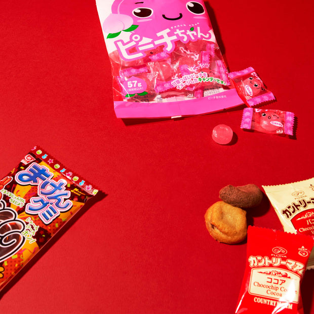 Experience the diverse flavors of Japan with the Dagashi Snack Crate (FBA) – a Japanese Snack Variety Box brimming with candy, chocolate, cookies, mochi, and ramune treats. Inside, you’ll find a pink peach-flavored candy bag, red cocoa cookie packets, and a brown snack packet—all artfully arranged on a vibrant red background. This crate is perfect for fans of authentic Japanese confections or anyone eager to explore the unique delights tucked into every Dagashi Snack Crate.