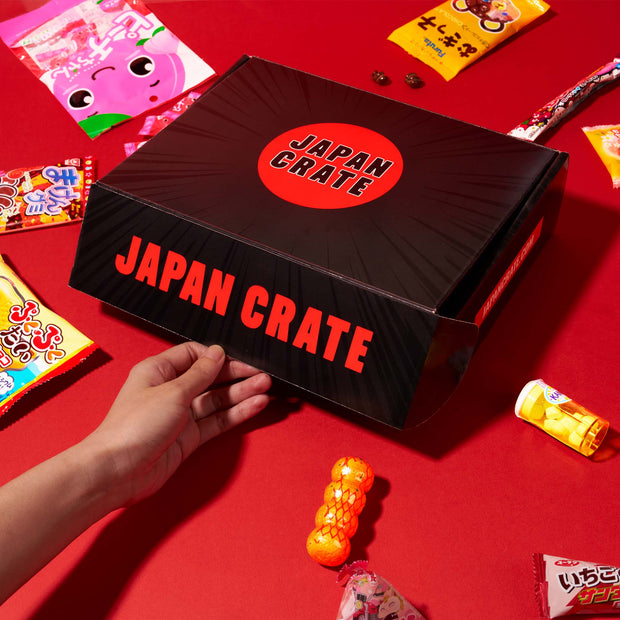 A hand holds a black box labeled “Japan Crate” on a red surface, surrounded by an array of vibrant Japanese snack and candy packages, showcasing the delightful assortment found in the Dagashi Snack Crate (FBA) - Japanese Snack Variety Box with Candy, Chocolate, Cookies, Mochi & Ramune Treats.