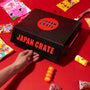 A hand holds a black box labeled “Japan Crate” on a red surface, surrounded by an array of vibrant Japanese snack and candy packages, showcasing the delightful assortment found in the Dagashi Snack Crate (FBA) - Japanese Snack Variety Box with Candy, Chocolate, Cookies, Mochi &amp; Ramune Treats. thumbnail 10