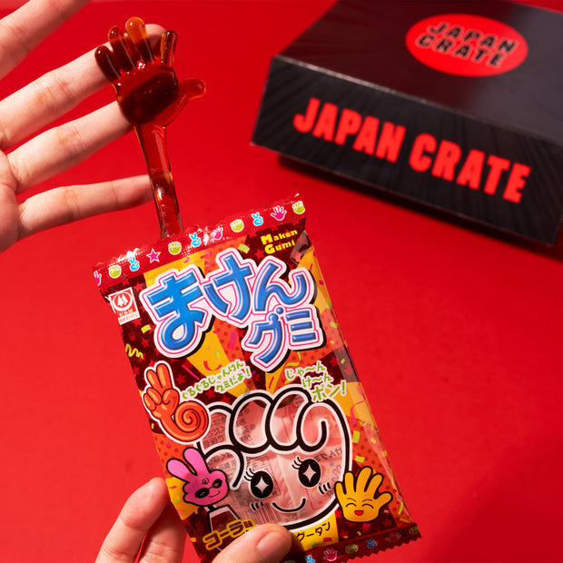 A hand holds a red hand-shaped gummy candy above colorful Japanese candy packaging, with a black Dagashi Snack Crate (FBA) – Japanese Snack Variety Box featuring candy, chocolate, cookies, mochi, and Ramune treats – displayed in the background on a red surface; an exciting preview of the unique snacks you’ll find inside every crate.