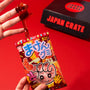 A hand holds a red hand-shaped gummy candy above colorful Japanese candy packaging, with a black Dagashi Snack Crate (FBA) – Japanese Snack Variety Box featuring candy, chocolate, cookies, mochi, and Ramune treats – displayed in the background on a red surface; an exciting preview of the unique snacks you’ll find inside every crate. thumbnail 2