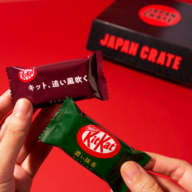 Two hands hold Japanese Kit Kat bars, one in a dark red wrapper and the other in a green wrapper, with a black Dagashi Snack Crate (FBA) - Japanese Snack Variety Box with Candy, Chocolate, Cookies, Mochi & Ramune Treats filled with Japanese snacks in the background on a red surface.