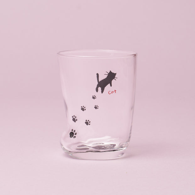 Introducing The Cat Lover’s Box: a clear drinking glass featuring a slight dent, adorned with black cat paw prints and a small black cat graphic with “cat” written in red beside it, set against a light pink background—perfect for serving your favorite cat-themed snacks or as a thoughtful addition to any Japanese cat gifts collection.