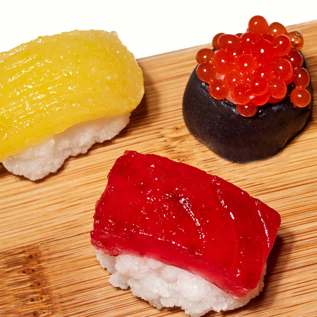 Three exquisite pieces of sushi rest on a wooden board: yellow mango nigiri, red tuna nigiri, and black caviar gunkan. Nearby, a DIY Candy Kit Pack (6-Pack) by Bokksu Market offers a playful twist, inspiring the creation of homemade candies that mirror these delightful treats.