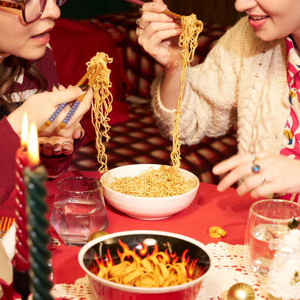 Two people enjoy noodles with chopsticks from the Spicy Challenge Crate, inspired by Japan-exclusive spicy snacks. Glasses of water and candles sit on a red tablecloth, enhancing the bold flavors of this unique crate.