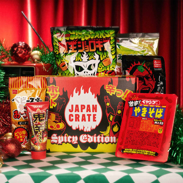 A Spicy Challenge Crate is surrounded by fiery Japanese snacks and instant noodles, including Japan-exclusive spicy treats, all displayed on a green and white checkered tablecloth with festive decorations.