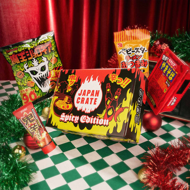 The Spicy Challenge Crate, featuring a selection of spicy Japanese snacks, is displayed on a checkered table with festive decorations and a red curtain in the background.