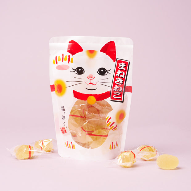 A white snack bag featuring a maneki-neko (beckoning cat) design, filled with individually wrapped yellow candies, makes a charming addition to The Cat Lover’s Box or a delightful Japanese cat gift, displayed against a light pink background.