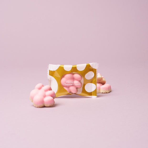 A pink paw-shaped chocolate treat in a clear and gold polka dot wrapper, with two unwrapped chocolates in the background on a light pink surface—perfect as a sweet addition to The Cat Lover’s Box or for cat-themed snacks.