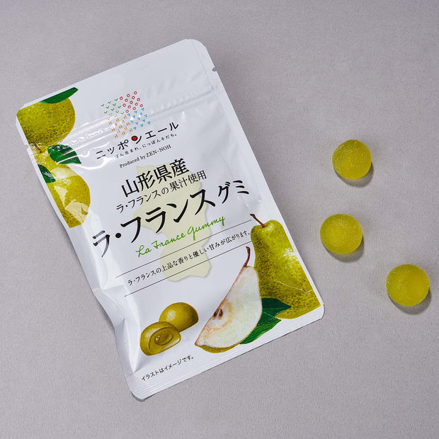 A white box labeled Japanese Fruits Gummy Box, featuring La France pear flavor, sits on a gray surface with three round yellow-green gummies displayed to its right.