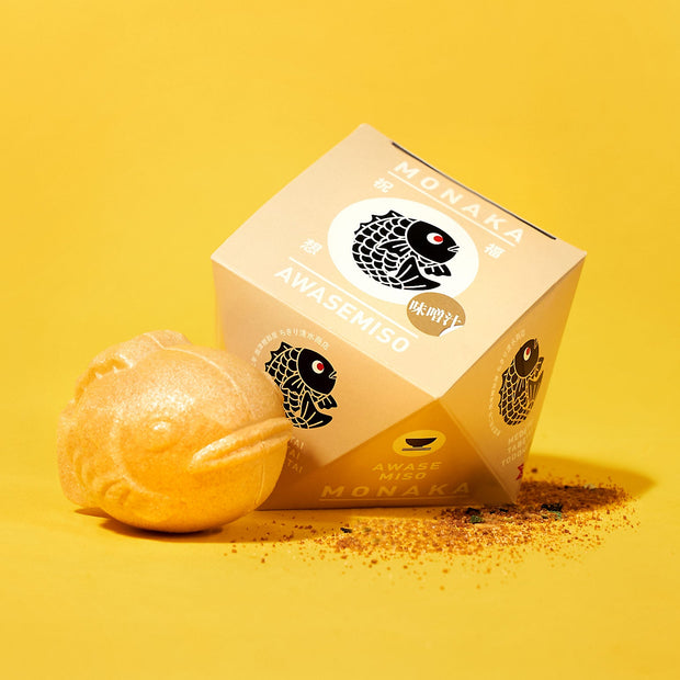 A fish-shaped monaka snack from The Japanese Savory Snack Box sits beside its beige, cube-shaped Awase Miso Monaka packaging on a yellow background with scattered seasoning.