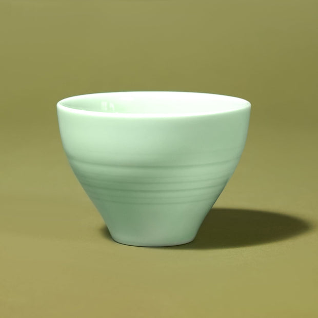 The Japanese Tea Box features a pale green ceramic cup with a smooth, matte finish, ideal for enjoying matcha. It rests on a plain olive-green surface, complemented by a matching background.