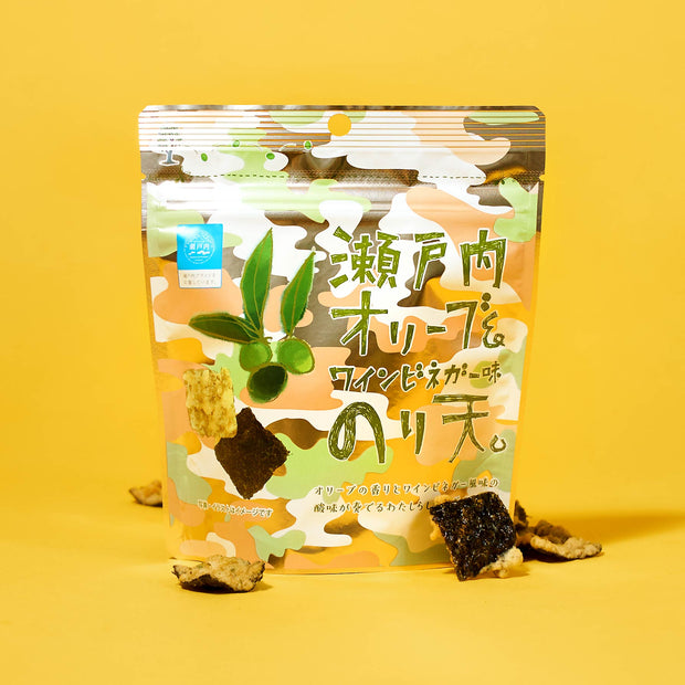 The Japanese Savory Snack Box, featuring a camouflage-patterned package with bold text, stands upright on a yellow background, surrounded by scattered pieces of seaweed snack.