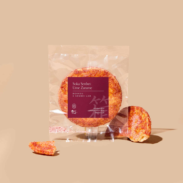 A round senbei rice cracker from Bokksu’s Japanese Savory Snack Box is shown in clear packaging labeled "Soka Senbei Ume Zarame" on a beige background, with a broken piece and crumbs beside it.
