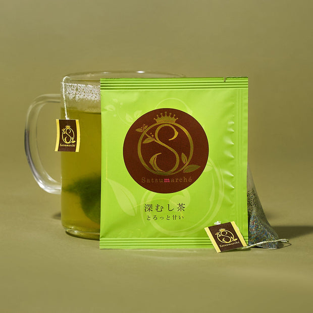 A packet labeled "The Japanese Tea Box" stands in front of a glass mug of matcha tea with a tea bag and tag, set against a plain olive background.