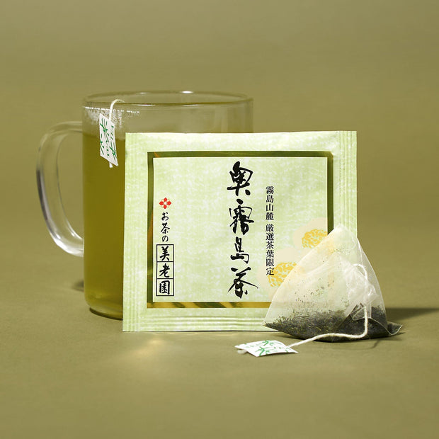 A glass mug of green tea sits beside The Japanese Tea Box and a used pyramid-shaped tea bag on a green surface with a green background.
