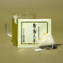 A glass mug of green tea sits beside The Japanese Tea Box and a used pyramid-shaped tea bag on a green surface with a green background. thumbnail 5