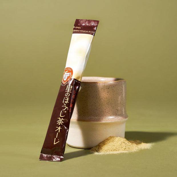 The Japanese Tea Box includes a ceramic cup, a matcha powder stick packet with Japanese text, and a small pile of matcha drink mix displayed on a light green surface.