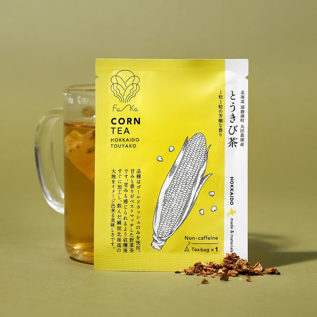 A glass mug of corn tea beside a yellow "The Japanese Tea Box" packet with corn illustration, loose tea leaves scattered nearby, evoking the tradition of Japanese tea boxes.