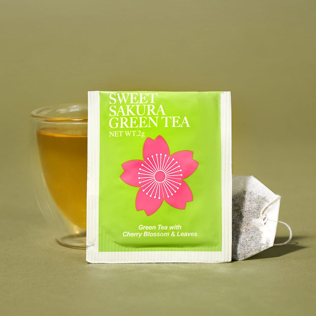 A glass cup of green tea sits behind The Japanese Tea Box, with a tea bag and string beside it, on a plain light brown background.