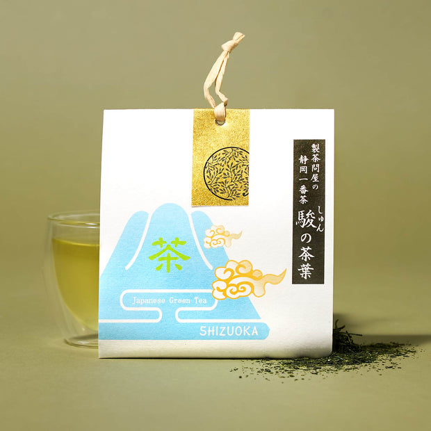 The Japanese Tea Box, with a blue Mount Fuji design, gold label, and Japanese text, is displayed next to a glass of Shizuoka green tea and loose leaves on a light green background.