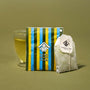 A glass cup of green tea sits behind The Japanese Tea Box and an unused tea bag on a plain olive background. thumbnail 12