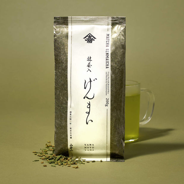 The Japanese Tea Box with Matcha Iri Genmaicha loose green tea leaves scattered beside a glass mug of vibrant green tea, all set on a yellow background.