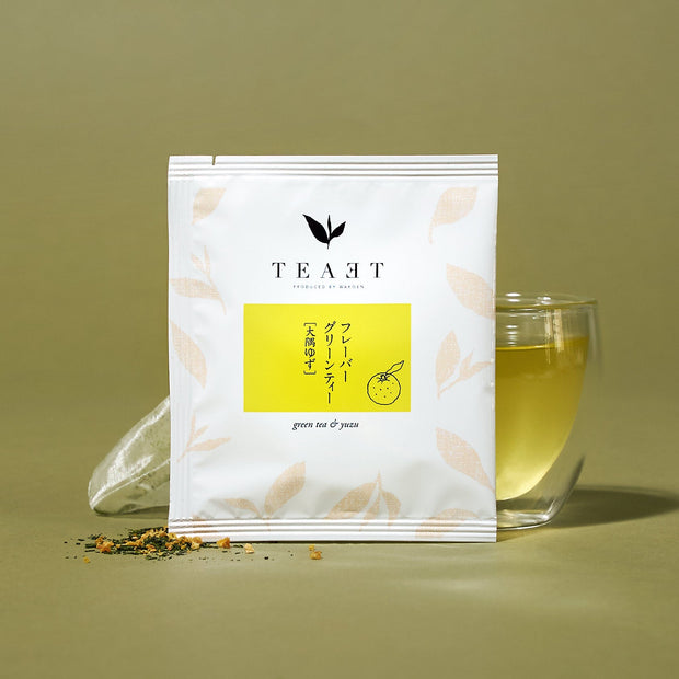 A packet of The Japanese Tea Box green tea & yuzu stands before a glass mug of light yellow tea, with loose tea leaves scattered nearby.