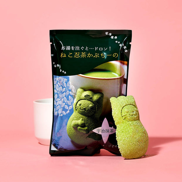 The Cat Lover's Box by Bokksu features green tea snacks shaped like cats, plus a matching cat-shaped treat—ideal for Manekineko Jelly fans—all beautifully displayed against a pink background.