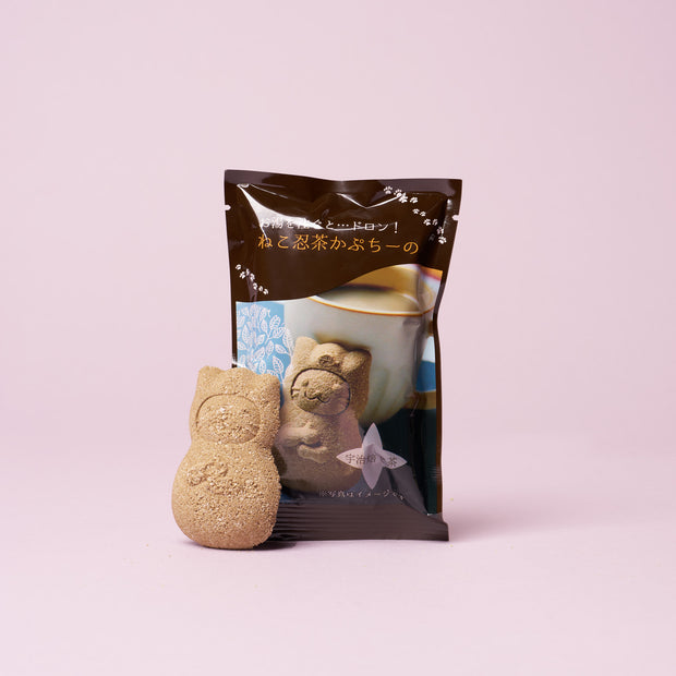 A cat-shaped brown sugar candy from The Cat Lover’s Box stands in front of a package featuring a cup of tea with Japanese text, set against a light pink background—making it the perfect treat for fans of cat-themed snacks and unique Japanese cat gifts.