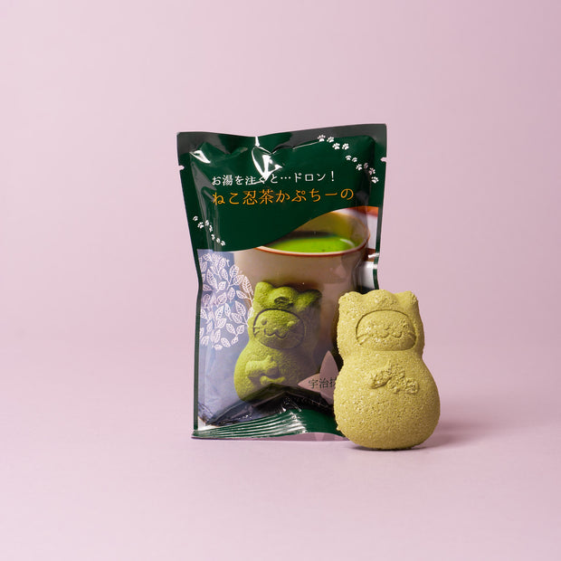 A green, cat-shaped powdered candy sits in front of its packaging featuring a cup of green tea on a pink background—an ideal treat for The Cat Lover’s Box subscribers seeking unique cat-themed snacks from Japan.
