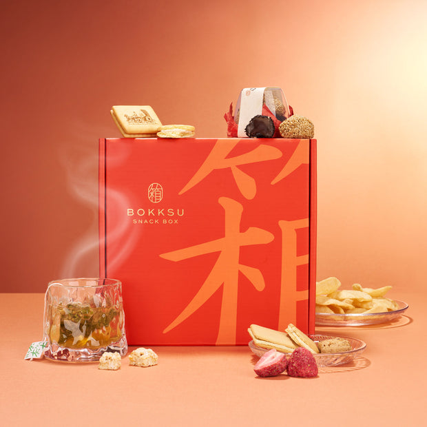 A Bokksu Snack Box: Seasons of Japan sits among assorted Japanese snacks and a steaming glass of tea, inviting you to experience Japan's flavors against a warm orange background.
