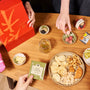 A tabletop with the Bokksu Snack Box: Seasons of Japan, a glass of tea, and two people reaching for treats—one holding a green tea packet, the other a red box—captures the authentic taste of Japan. thumbnail 2