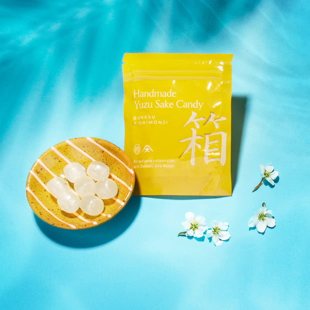 A Bokksu Snack Box: Seasons of Japan pouch labeled "Handmade Yuzu Sake Candy" sits by round candies and white blossoms on a blue surface—offering an authentic taste of Japan in each delightful snack.