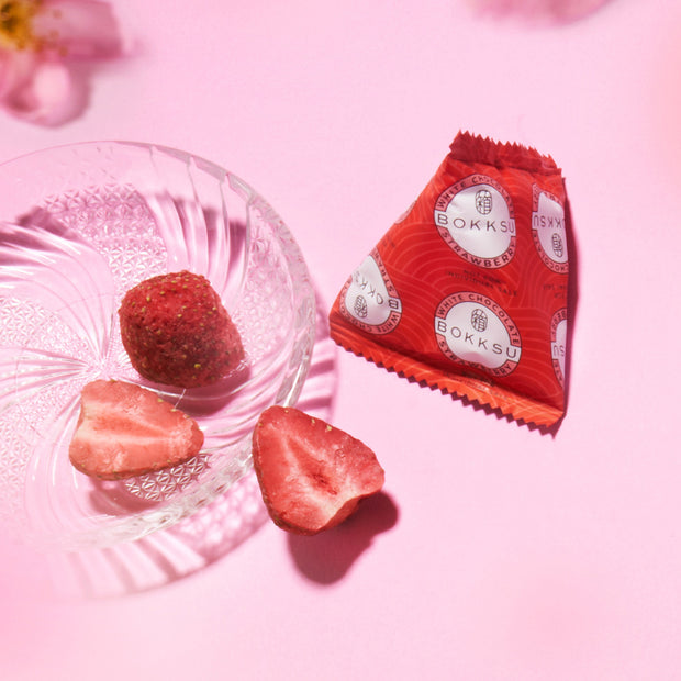 A clear glass dish with two freeze-dried strawberries and one coated strawberry sits beside a red Bokksu Snack Box: Seasons of Japan wrapper on a pink background, offering an authentic taste of Japanese snacks.