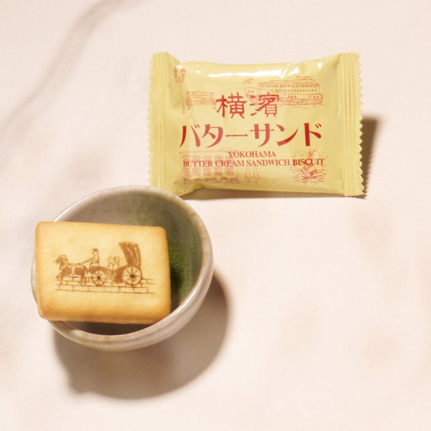 A butter cream sandwich biscuit with a horse-drawn carriage design sits in a small bowl beside its yellow packaging labeled "Yokohama Butter Cream Sandwich Biscuit," featured in the Bokksu Snack Box: Seasons of Japan for an authentic Japanese treat.