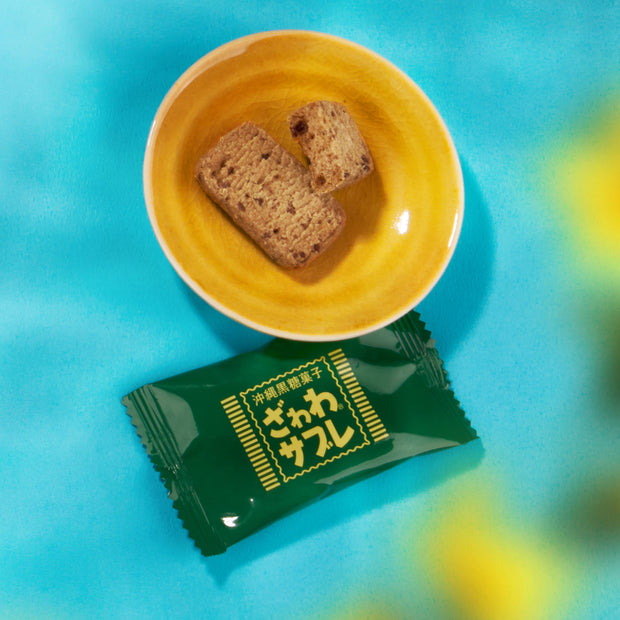 A yellow bowl with two rectangular cookies rests above a green, Japanese-labeled packet on a blue and yellow surface—offering an inviting peek into the Bokksu Snack Box: Seasons of Japan experience.
