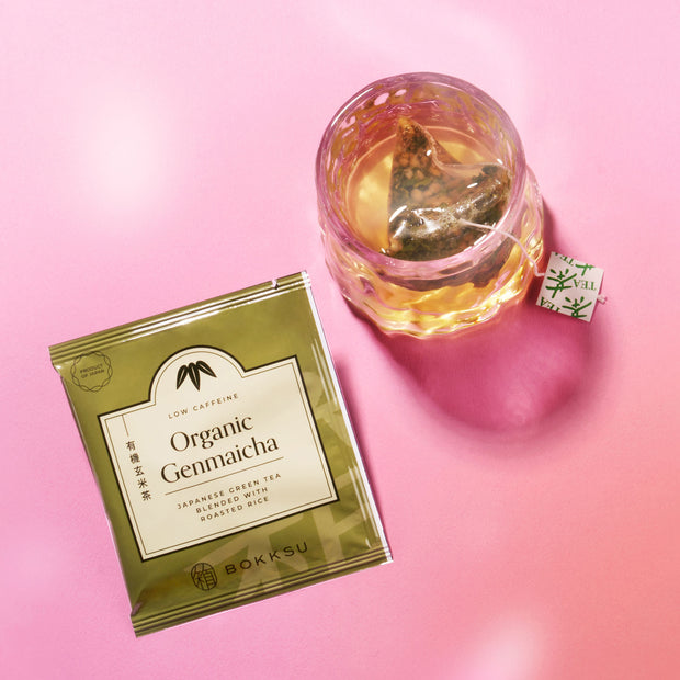 A packet of Organic Genmaicha Japanese green tea and a glass cup with tea sit on a pink surface, perfect for enjoying alongside the Bokksu Snack Box: Seasons of Japan.