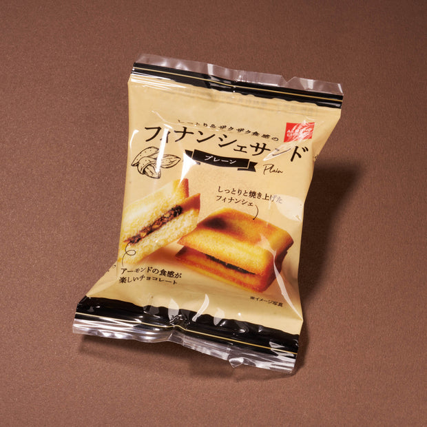 Handpicked in Japan and featured in April '26 Bokksu, "Financier Sand Plain" is a Japanese snack shown on a brown surface, with its beige wrapper displaying an image of a filled cake.